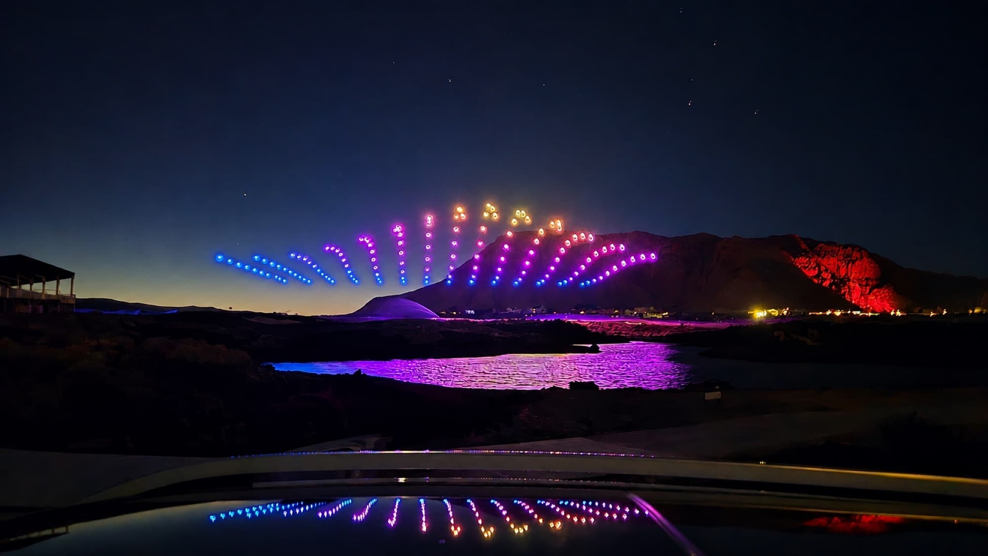 A wide fan formation in drones above a resort lake, mirrored on the water below