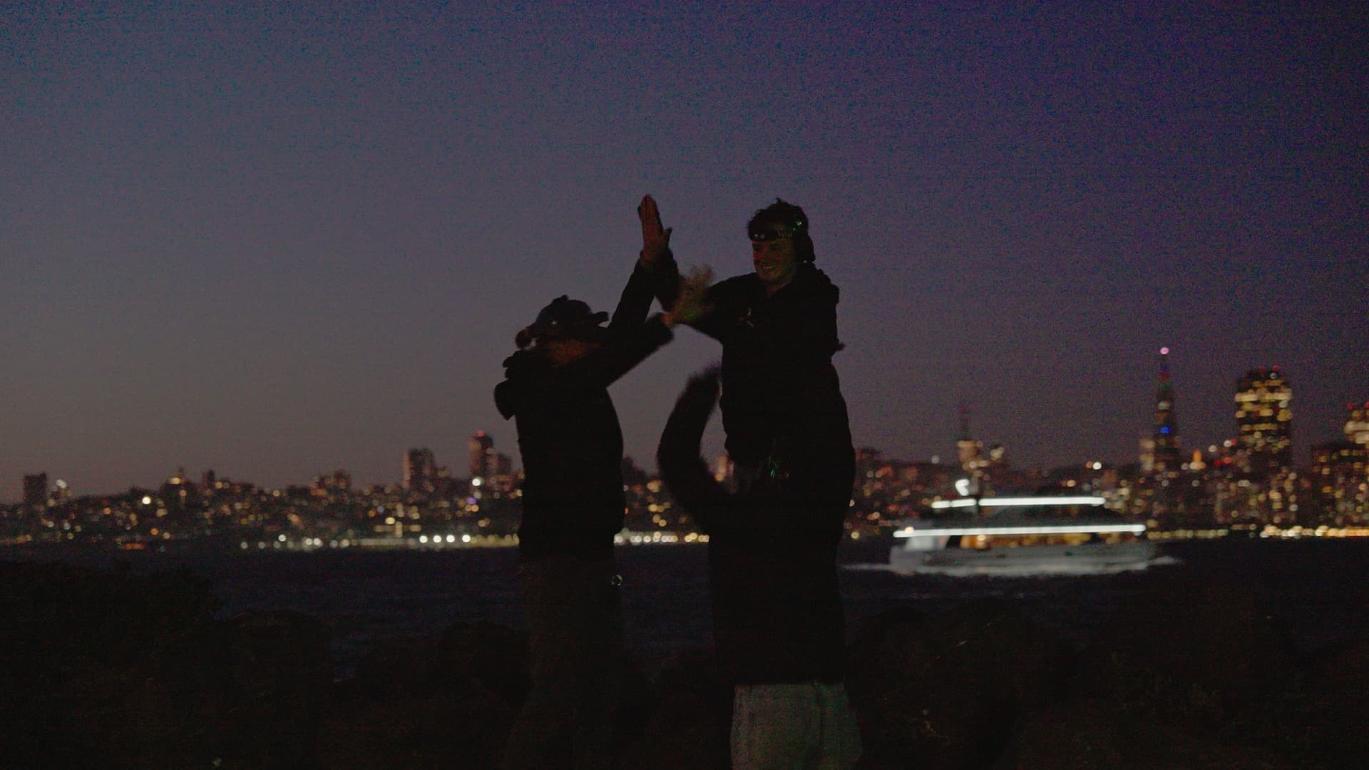 Two crew members high-fiving at dusk on the launch site, the San Francisco skyline lit behind them