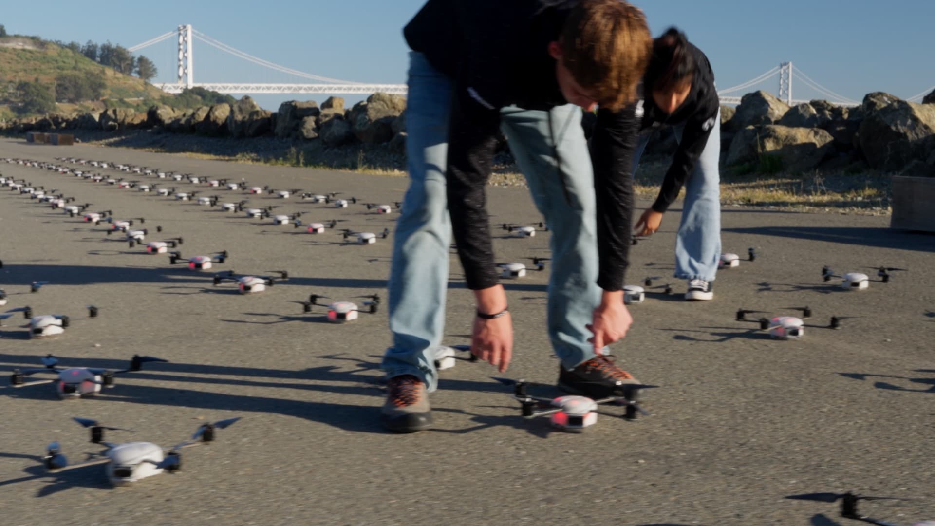 Two crew members placing drones in a launch grid on asphalt, Bay Bridge in the distance