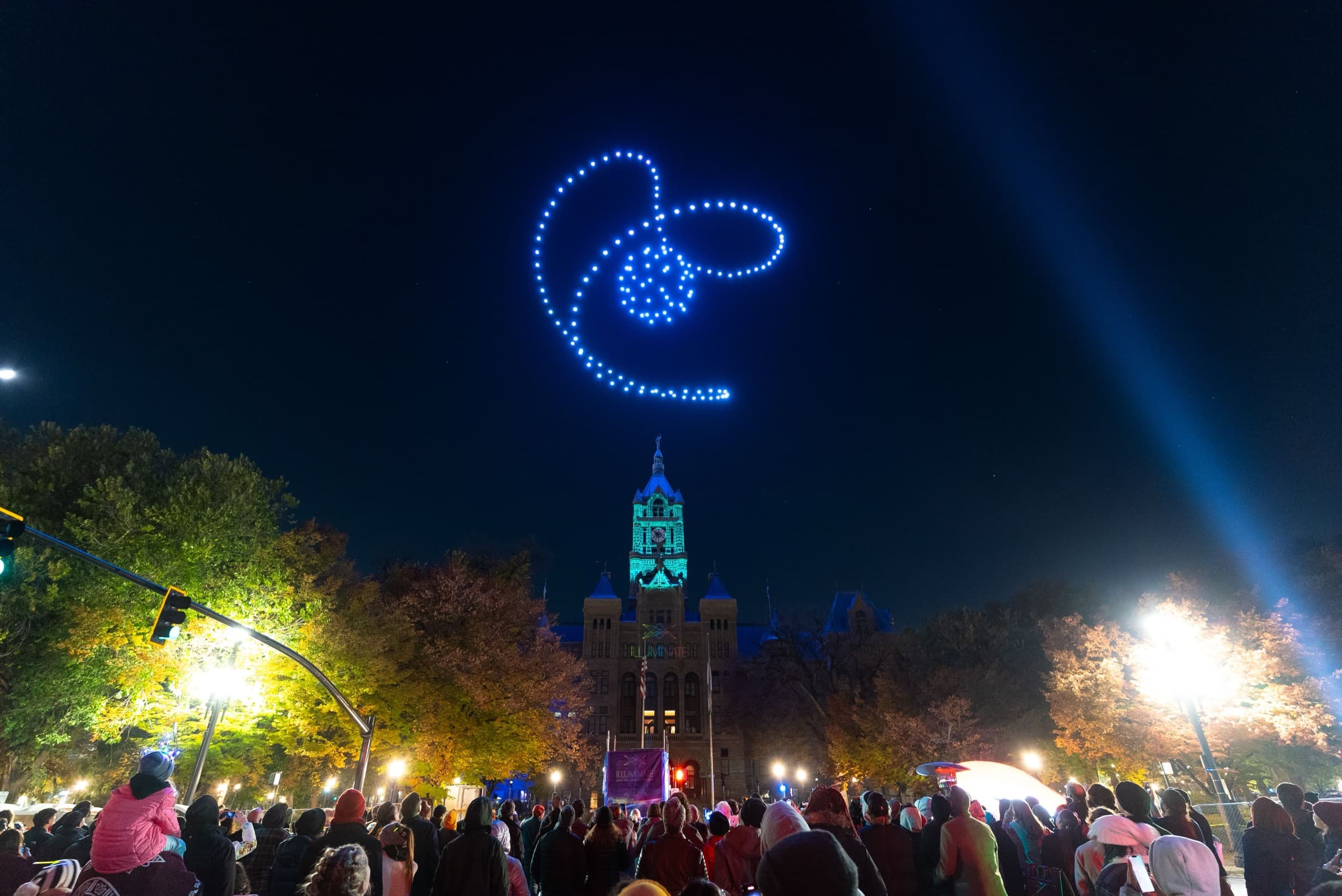 A crowd watching a drone formation above a lit civic building