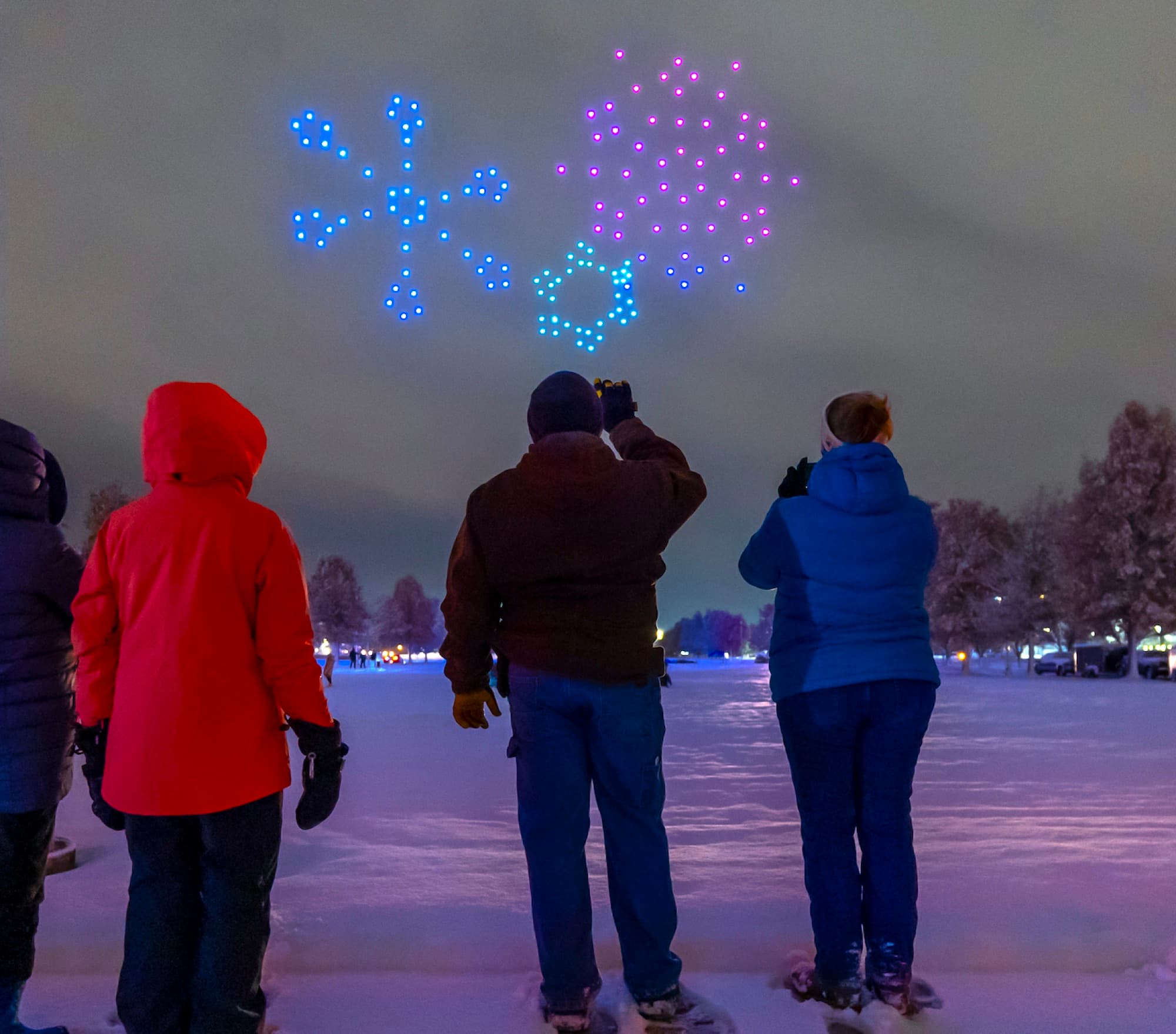 Three people in winter coats watching snowflake formations in drones over a snow-covered park