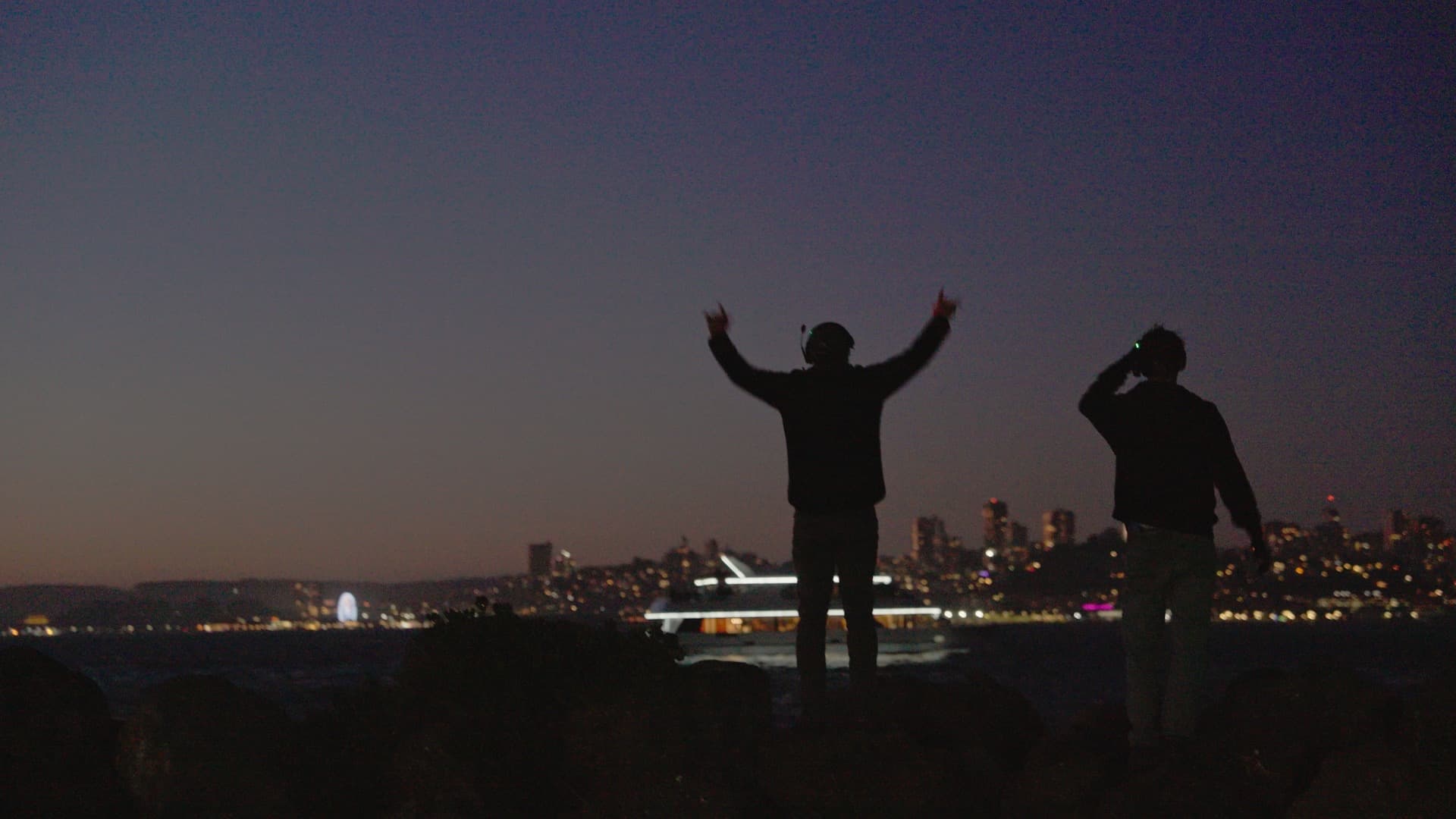 Two spectators silhouetted against a drone show over the water at dusk